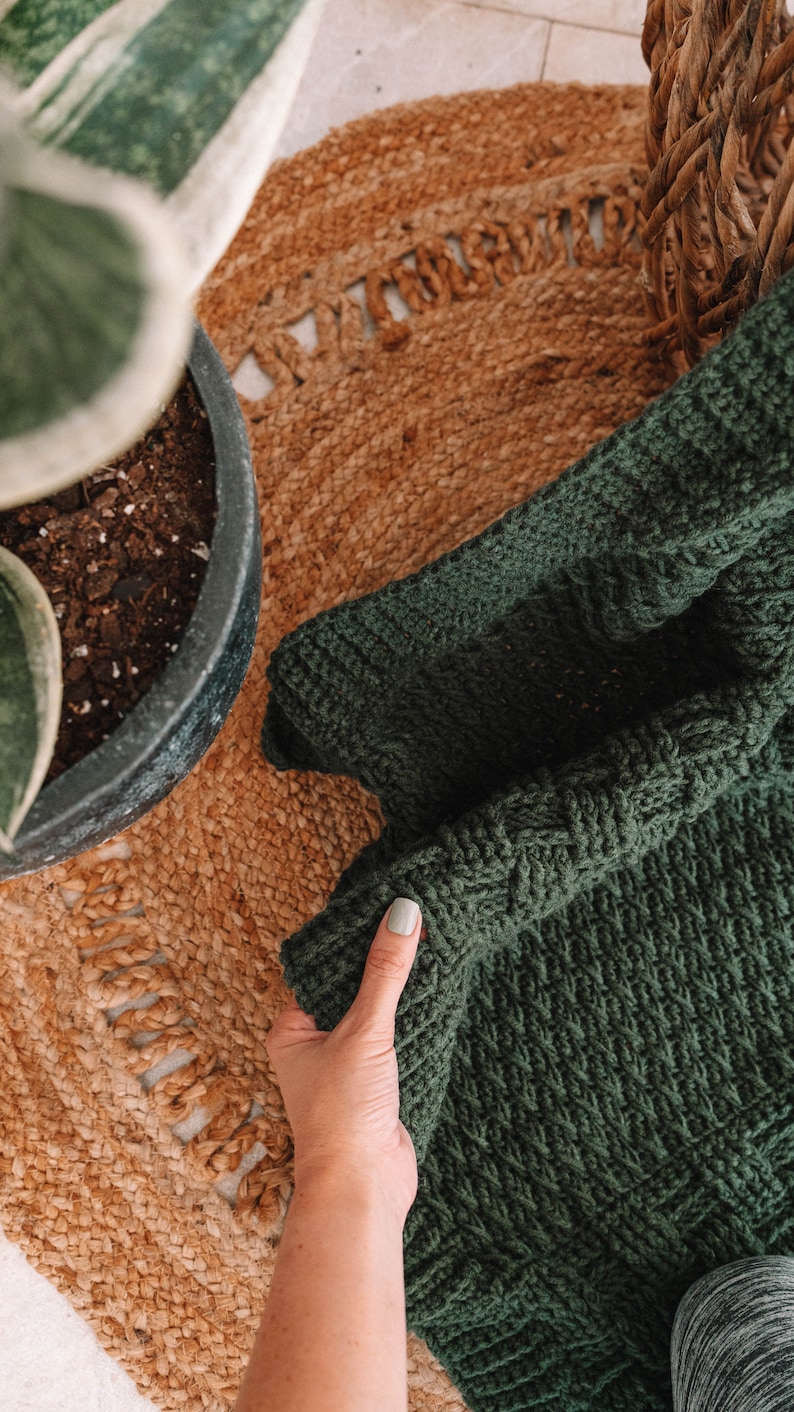 Puede incluir: Una manta de ganchillo de color verde bosque oscuro con un patr&oacute;n de punto de cester&iacute;a denso y texturizado. La tela es gruesa y uniforme, mostrando una construcci&oacute;n de hilo consistente en toda la pieza, destacando los intrincados bucles entrelazados.