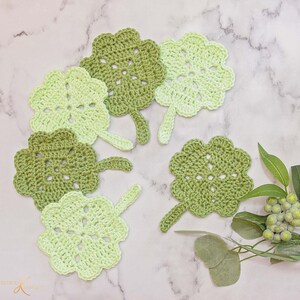 Handmade green crochet coasters designed to resemble leaves placed on a marble surface, with a sprig of green berries and leaves to the side, making an exceptional handmade gift.