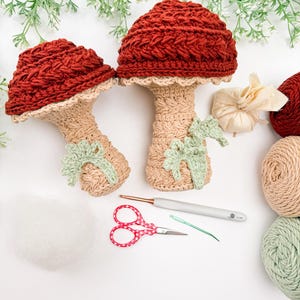 May include: Two crocheted mushroom figures with dark red caps and beige stems, each featuring a small light green leaf detail. The set includes a silver crochet hook, red handled scissors, a green sewing needle, and yarn balls in red, beige, and green.