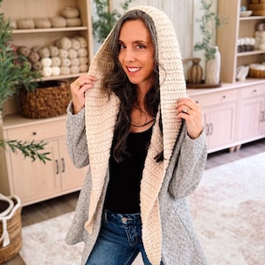 May include: A hooded cardigan featuring a textured, cream-colored crocheted hood and front panels, contrasted with a grey, herringbone-patterned knit body. The garment is worn over a black top and blue denim jeans, showcasing a long-sleeved design.