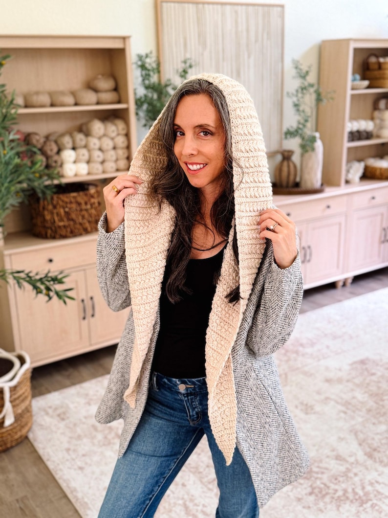 May include: A hooded cardigan featuring a textured, cream-colored crocheted hood and front panels, contrasted with a grey, herringbone-patterned knit body. The garment is worn over a black top and blue denim jeans, showcasing a long-sleeved design.