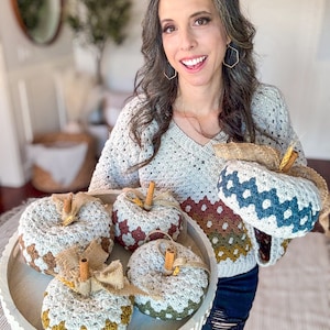 Pode incluir: Um conjunto de cinco abóboras feitas à mão em crochê com padrão granny square em vários tons terrosos, incluindo creme, castanho, verde-azeitona e azul-petróleo escuro. Cada abóbora tem um caule de pau de canela e um laço de fita de juta.