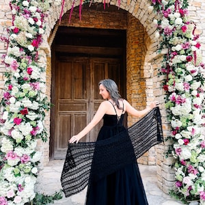 A woman in a black dress stands in front of a large wooden doorway framed by a floral arch made of white and pink flowers, holding out a black shawl behind her, perfectly accessorized with unique jewellery.