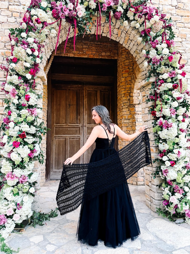 A woman in a black dress stands in front of a large wooden doorway framed by a floral arch made of white and pink flowers, holding out a black shawl behind her, perfectly accessorized with unique jewellery.