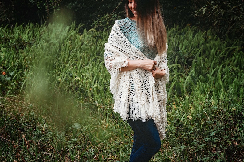 A woman wearing a white shawl decorated with the intricate crochet pineapple stitch, with fringed edges. She stands in an outdoor green space, surrounded by greenery and sunshine.