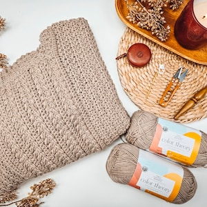 Knitted sweater laid on a white surface surrounded by yarn skeins, knitting tools, a red candle, and dried flowers on a woven mat—a perfect handmade gift reminiscent of vintage clothes.