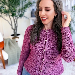 May include: A close-up of a purple crochet cardigan with pearl buttons. The cardigan has long sleeves and a textured stitch pattern. The person is wearing blue jeans. The background includes a green plant and a chair.