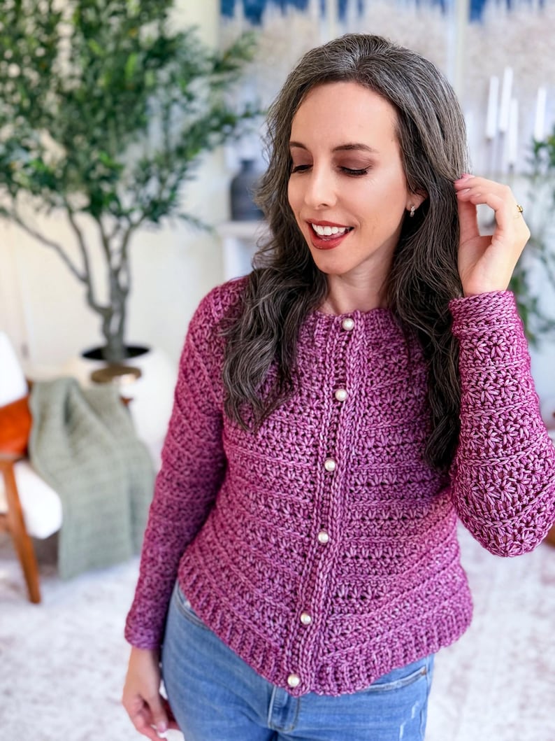 May include: A close-up of a purple crochet cardigan with pearl buttons. The cardigan has long sleeves and a textured stitch pattern. The person is wearing blue jeans. The background includes a green plant and a chair.