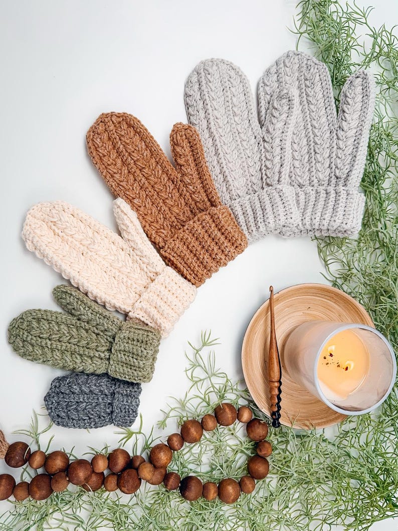 May include: Five pairs of crocheted mittens in various colours, including grey, beige, brown, green, and dark grey. The mittens are arranged in a semi-circle on a white surface. There is a wooden crochet hook, a wooden bead garland, and a candle on a wooden coaster in the background.