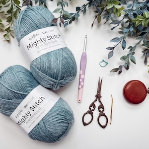 May include: A crafting still life featuring two balls of blue-grey yarn labeled "Mighty Stitch", a crochet hook, scissors, a tape measure, and other accessories. Greenery accents the composition, suggesting a creative hobby or craft project.
