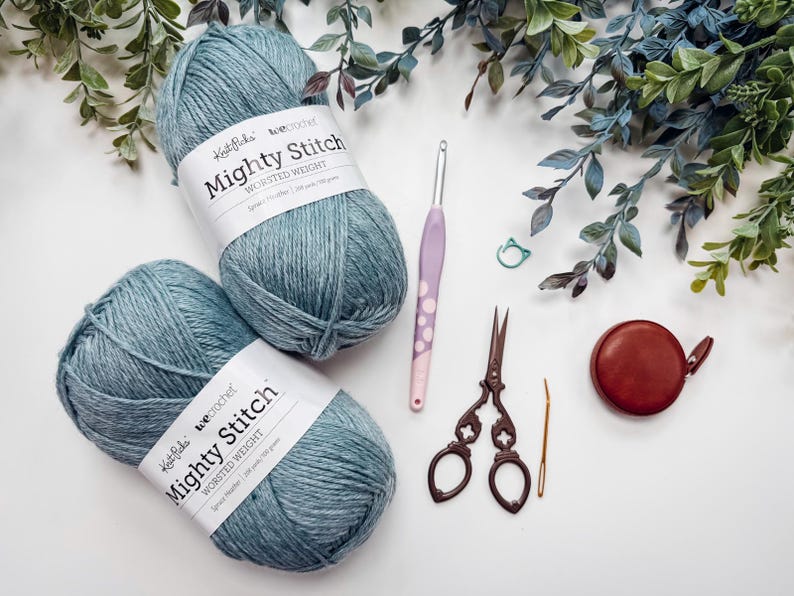 May include: A crafting still life featuring two balls of blue-grey yarn labeled "Mighty Stitch", a crochet hook, scissors, a tape measure, and other accessories. Greenery accents the composition, suggesting a creative hobby or craft project.