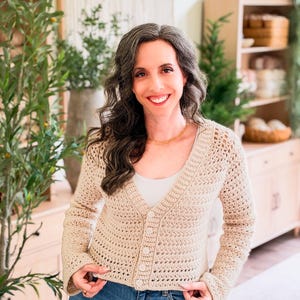 May include: A beige crochet cardigan featuring a V-neckline, long sleeves, and a front button closure with four matching buttons. The garment is crafted with a repeating horizontal stitch pattern and is worn over a plain white undershirt and blue jeans.