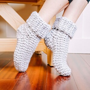 May include: A pair of grey knitted slipper socks with folded cuffs. The socks are being worn by a person sitting on a wooden bench.