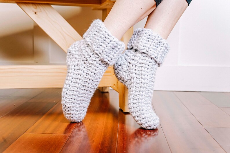 May include: A pair of grey knitted slipper socks with folded cuffs. The socks are being worn by a person sitting on a wooden bench.