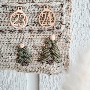 May include: Close-up of a handmade crochet advent calendar. The calendar is beige with brown flecks and features wooden ornaments with the numbers 23 and 24. Two green yarn Christmas trees are also attached to the calendar.