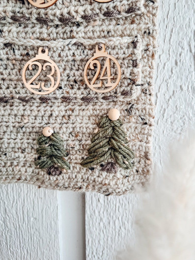 May include: Close-up of a handmade crochet advent calendar. The calendar is beige with brown flecks and features wooden ornaments with the numbers 23 and 24. Two green yarn Christmas trees are also attached to the calendar.
