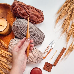 A hand holding a wooden crochet hook with various yarns, a candle, a measuring tape, scissors, and wheat stalks spread on a white surface could easily inspire handmade gifts or unique jewellery creations.