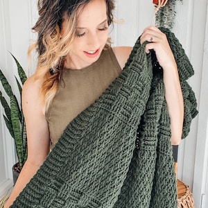 Puede incluir: Una manta tejida de color verde bosque oscuro que presenta una textura de tejido de cesta prominente. El textil est&aacute; elaborado con hilo grueso, creando un patr&oacute;n denso y uniforme de cuadrados entrelazados en toda la superficie de la tela.