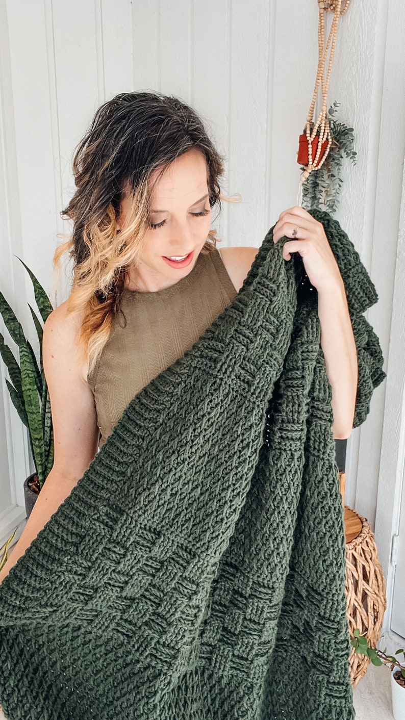Puede incluir: Una manta tejida de color verde bosque oscuro que presenta una textura de tejido de cesta prominente. El textil est&aacute; elaborado con hilo grueso, creando un patr&oacute;n denso y uniforme de cuadrados entrelazados en toda la superficie de la tela.