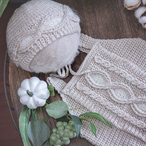 May include: A beige knitted baby hat and matching outfit with a cable knit design. The outfit has a button closure.