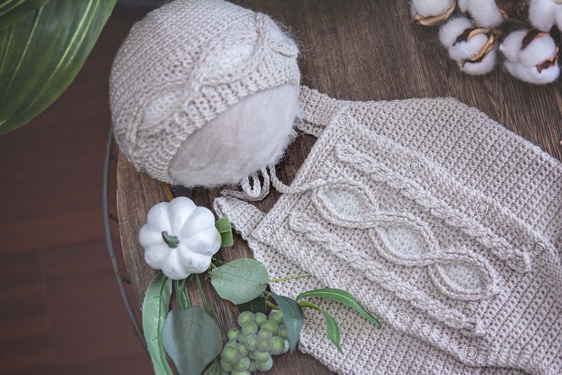May include: A beige knitted baby hat and matching outfit with a cable knit design. The outfit has a button closure.