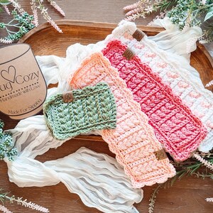 May include: A collection of handmade crochet mug cozies in various colors, including green, peach, pink, and white. Each cozy features a textured design and a small leather tag. The image also includes a cardboard tag that says "Mug Cozy".