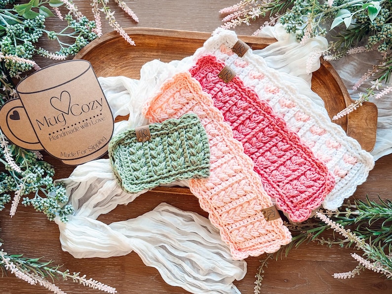 May include: A collection of handmade crochet mug cozies in various colors, including green, peach, pink, and white. Each cozy features a textured design and a small leather tag. The image also includes a cardboard tag that says "Mug Cozy".