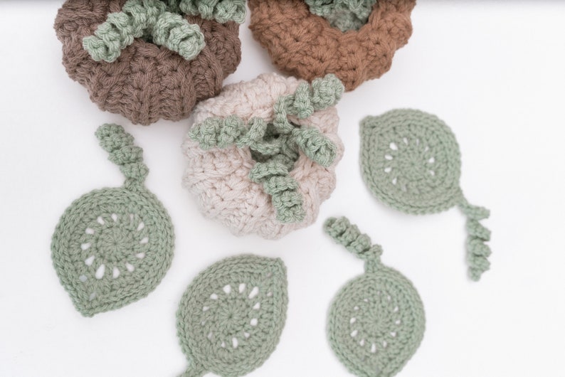 Three crochet pumpkins in taupe, tan and cream above five sage green leaf coasters with curly stems