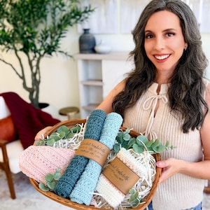 Pode incluir: Uma mulher segura uma cesta de vime cheia de panos de crochê feitos à mão em várias cores, incluindo azul, verde-azulado e rosa. Os panos estão enrolados e amarrados, com etiquetas que dizem "Handmade Washcloths with Love".