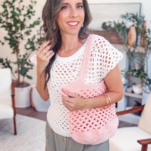 Woman in a white crochet top and gray skirt stands indoors, holding a pink crochet bag—a perfect handmade gift. There are plants and white chairs in the background.