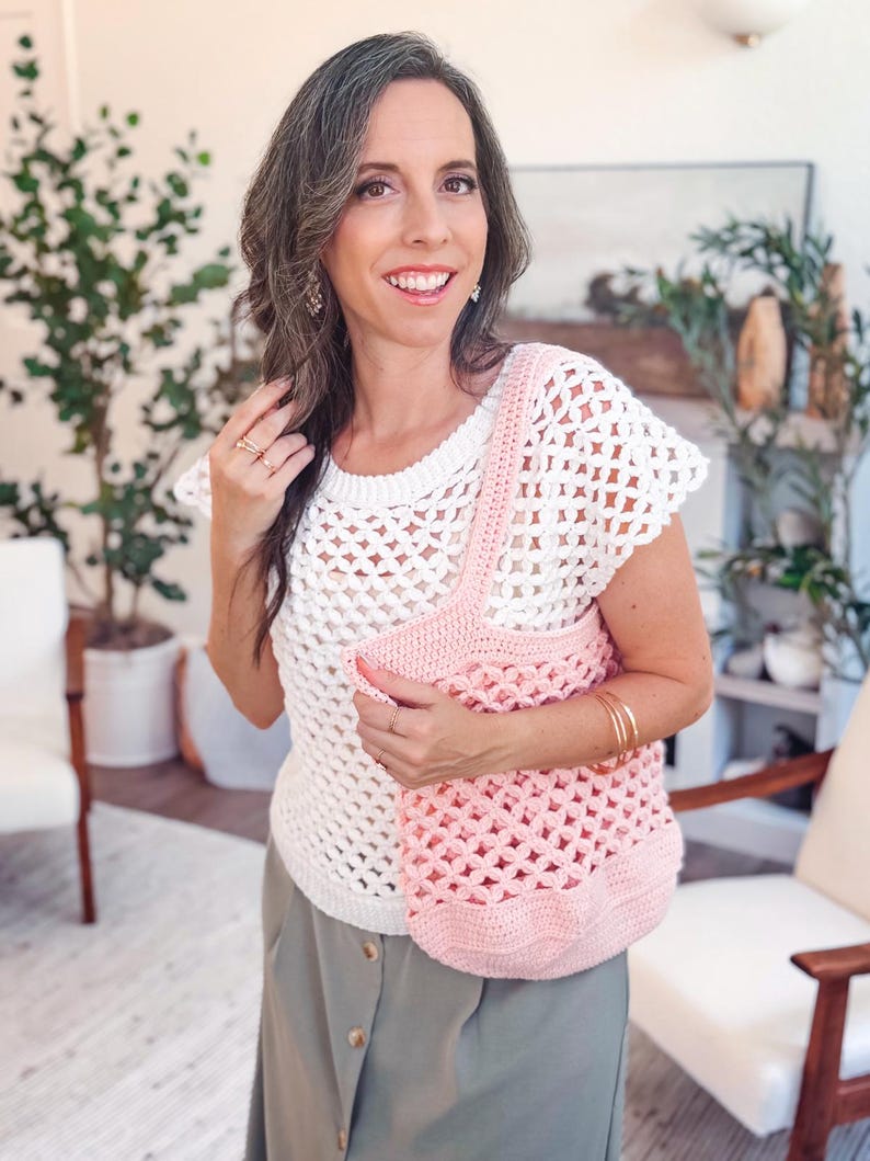 Woman in a white crochet top and gray skirt stands indoors, holding a pink crochet bag—a perfect handmade gift. There are plants and white chairs in the background.