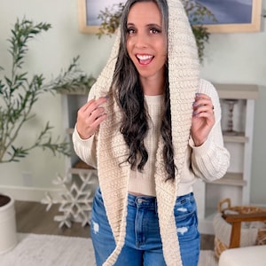 May include: Cream-coloured knitted hood with long scarf ends. The hood is made of a textured yarn and is worn over a cream-coloured sweater and blue jeans. A cosy accessory.