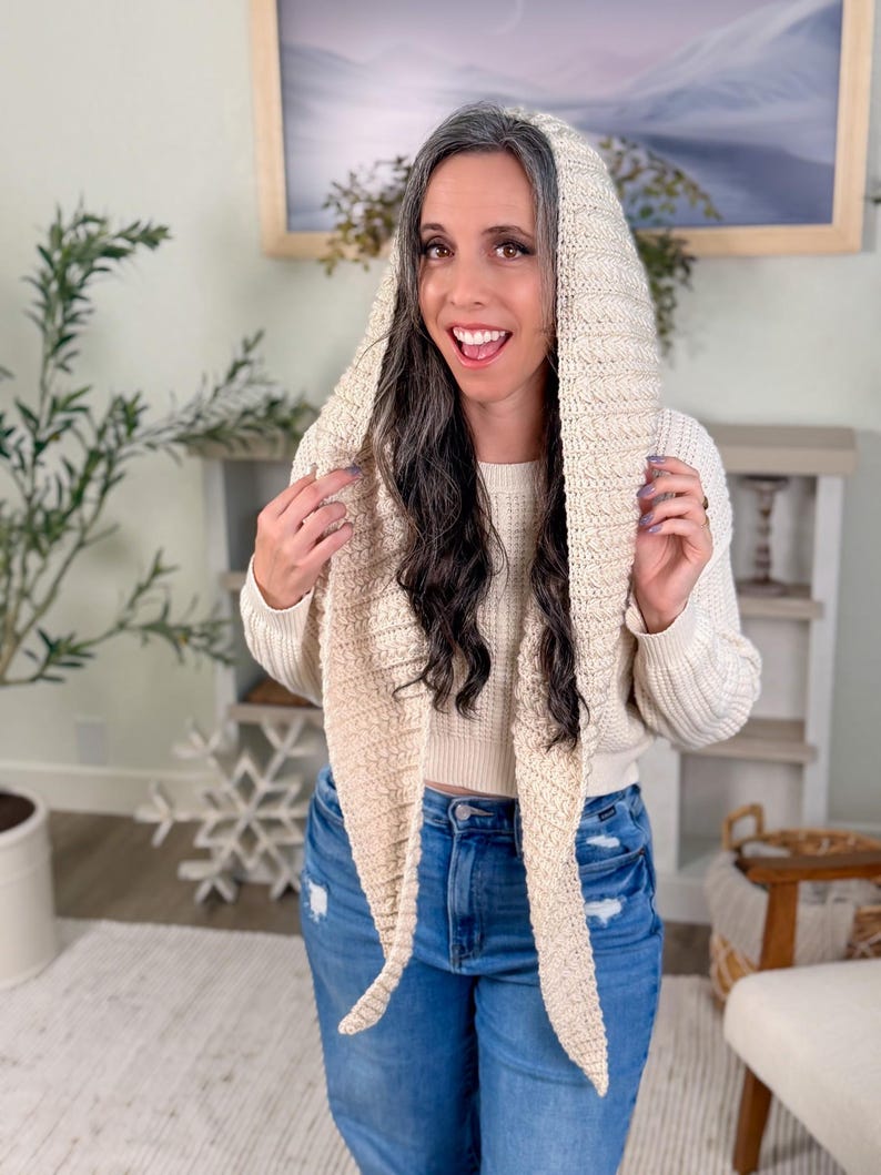 May include: Cream-coloured knitted hood with long scarf ends. The hood is made of a textured yarn and is worn over a cream-coloured sweater and blue jeans. A cosy accessory.