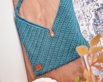 May include: A teal crochet hobo bag featuring a diagonal ribbed stitch pattern, a single braided shoulder strap, and a tan leather tab closure with a metal snap. A small square tan leather patch with the embossed letters QR is attached.