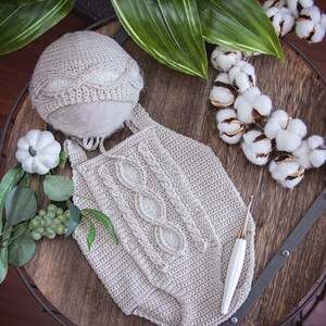 May include: A crocheted baby outfit with a cable knit pattern, a matching hat, and a crochet hook. The outfit is beige and the hat is white. The outfit is laid out on a wooden surface with a cotton wreath and greenery.