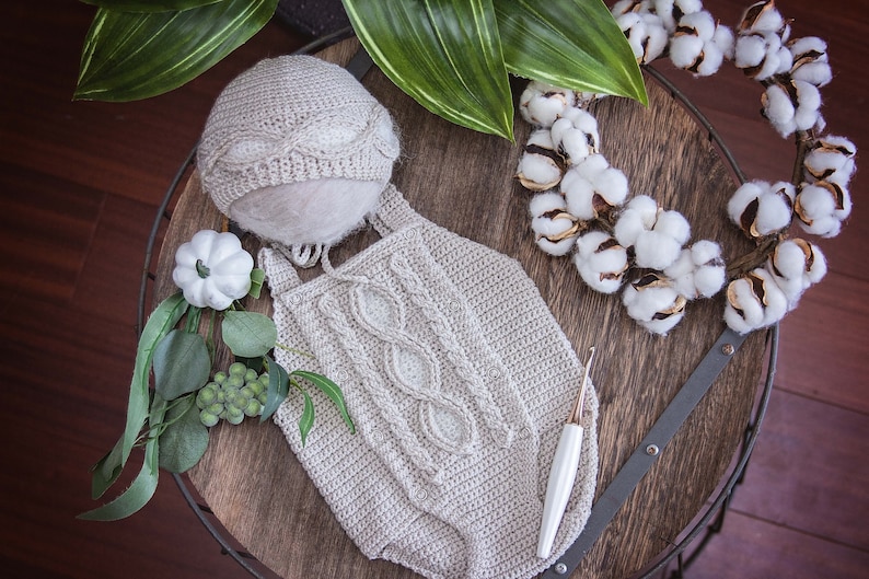 May include: A crocheted baby outfit with a cable knit pattern, a matching hat, and a crochet hook. The outfit is beige and the hat is white. The outfit is laid out on a wooden surface with a cotton wreath and greenery.