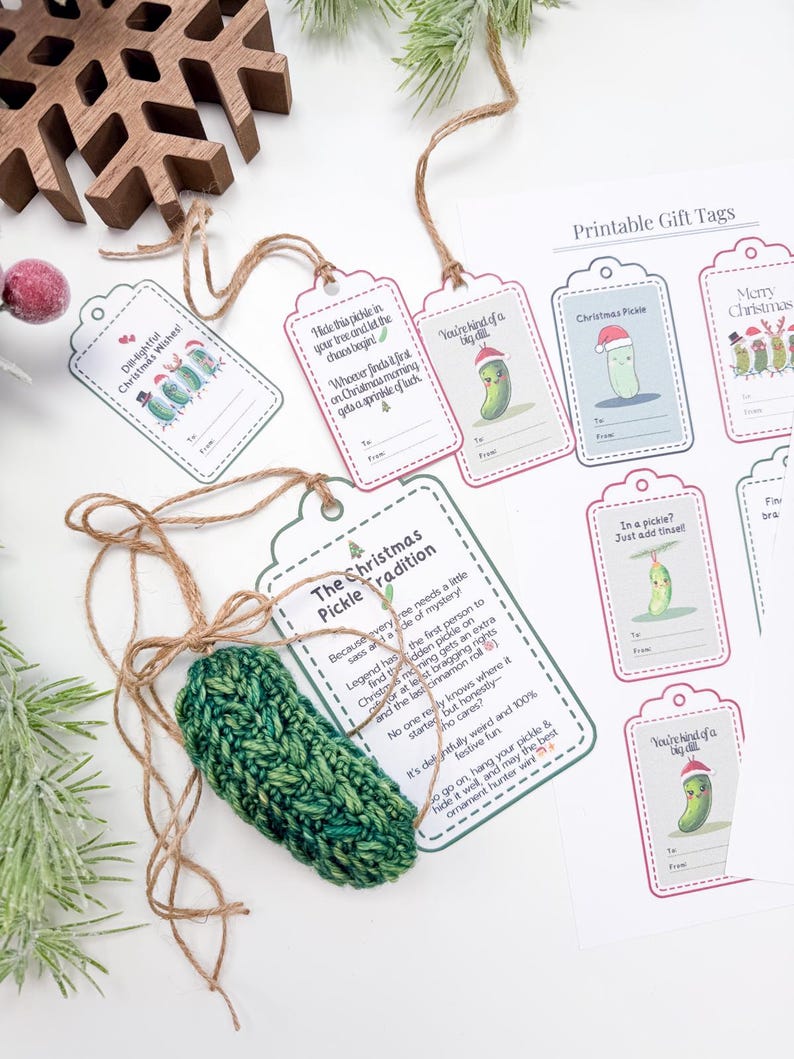 May include: A crocheted green pickle ornament with twine, accompanied by a sheet of printable gift tags featuring pickle illustrations and text including "The Christmas Pickle Tradition," "Dill-lightful Christmas Wishes," and "In a pickle?"