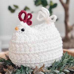 Puede incluir: Una figura de gallina tejida a ganchillo en color blanco con un patrón de puntos texturizados, una cresta de hilo rojo oscuro, un pequeño pico amarillo y ojos de cuentas negras. La gallina tiene una cola de bucles blancos y cuerpo redondo.