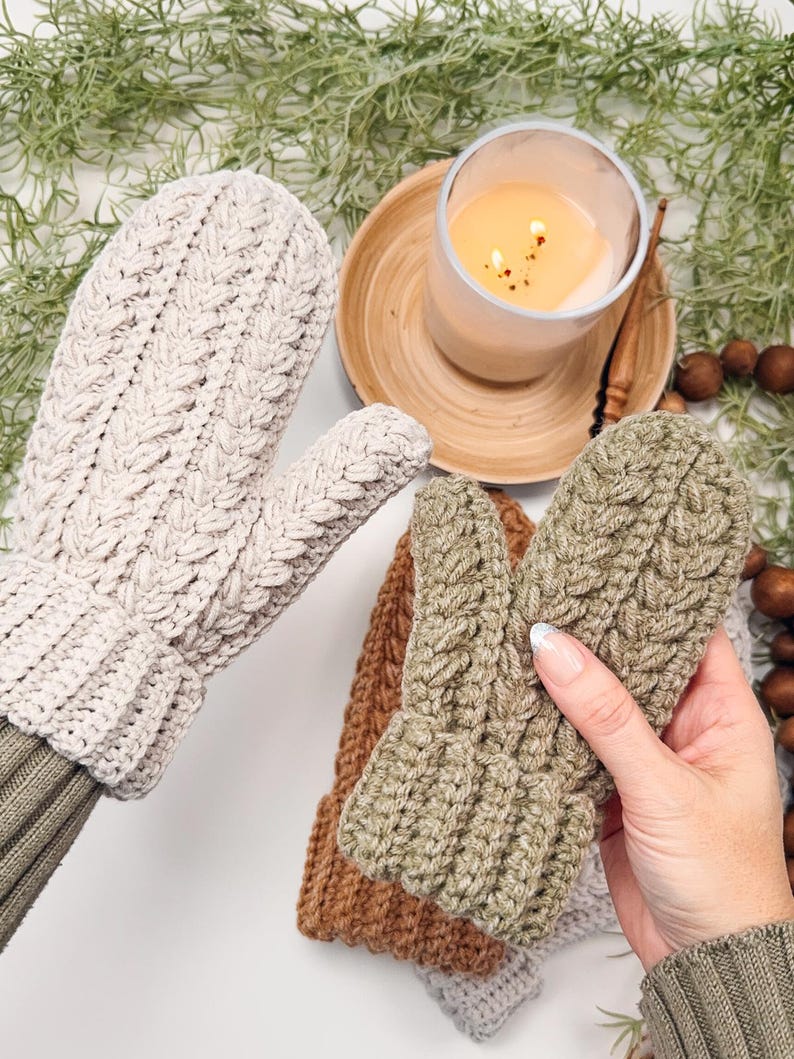 May include: Two pairs of knitted mittens, one in beige and one in green, with a cable knit pattern. The mittens are displayed on a white surface with a lit candle and greenery in the background.