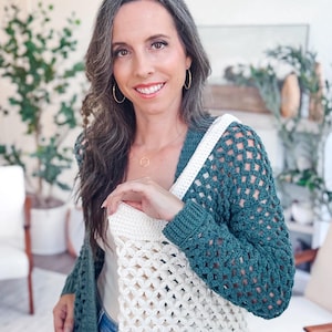 A woman with long brown hair wearing a green and white crocheted sweater stands indoors, smiling at the camera. Surrounded by plants and chairs, she looks as if she's ready to offer a handmade gift or showcase unique jewellery.