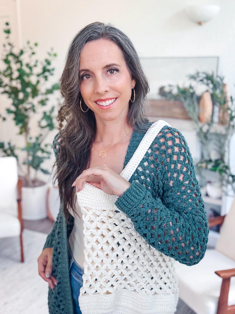 A woman with long brown hair wearing a green and white crocheted sweater stands indoors, smiling at the camera. Surrounded by plants and chairs, she looks as if she's ready to offer a handmade gift or showcase unique jewellery.