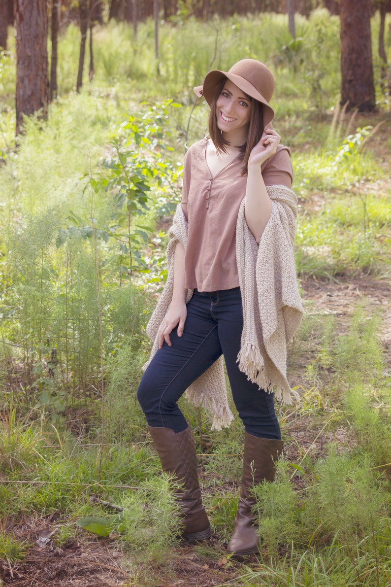 A woman wearing a cream crochet shawl wrap and hat, in a field of greenery.