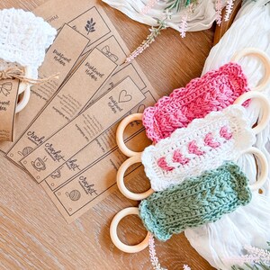 May include: A collection of handmade crochet dish towel holders in pink, white, and green, each attached to a wooden ring. The holders are displayed with kraft paper tags that read "Dish Towel Holder" and "crochet".