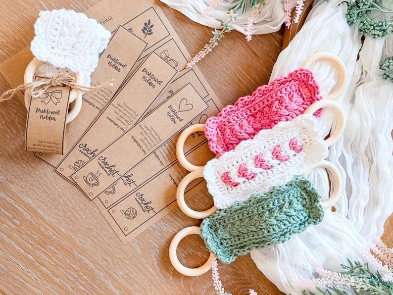 May include: A collection of handmade crochet dish towel holders in pink, white, and green, each attached to a wooden ring. The holders are displayed with kraft paper tags that read "Dish Towel Holder" and "crochet".