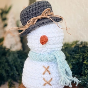 May include: A handmade crochet snowman with a gray hat tied with twine, an orange nose, and a light blue scarf. The snowman has brown stitched details and is a festive winter decoration.
