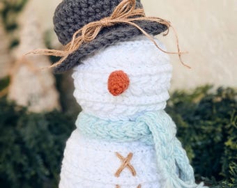 May include: A handmade crochet snowman with a gray hat tied with twine, an orange nose, and a light blue scarf. The snowman has brown stitched details and is a festive winter decoration.