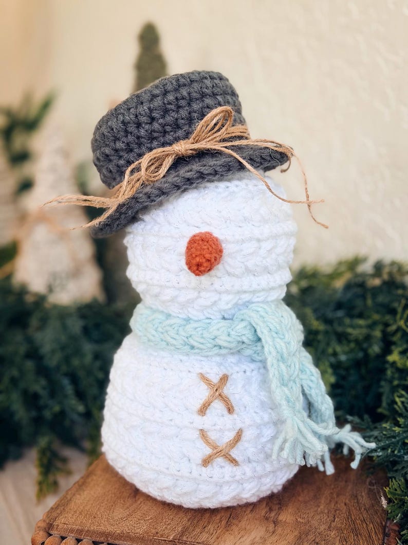 Close-up of a white textured crochet snowman with gray hat, carrot nose and light blue fringed scarf