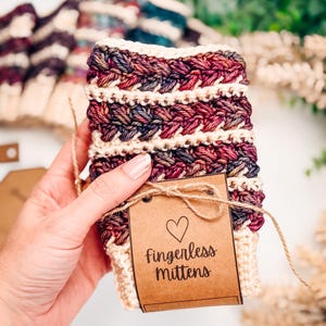 May include: Cream, burgundy, and purple fingerless mittens with a braided design. The mittens are handmade and have a brown tag that reads "Fingerless Mittens" with a heart symbol. The mittens are tied with twine.