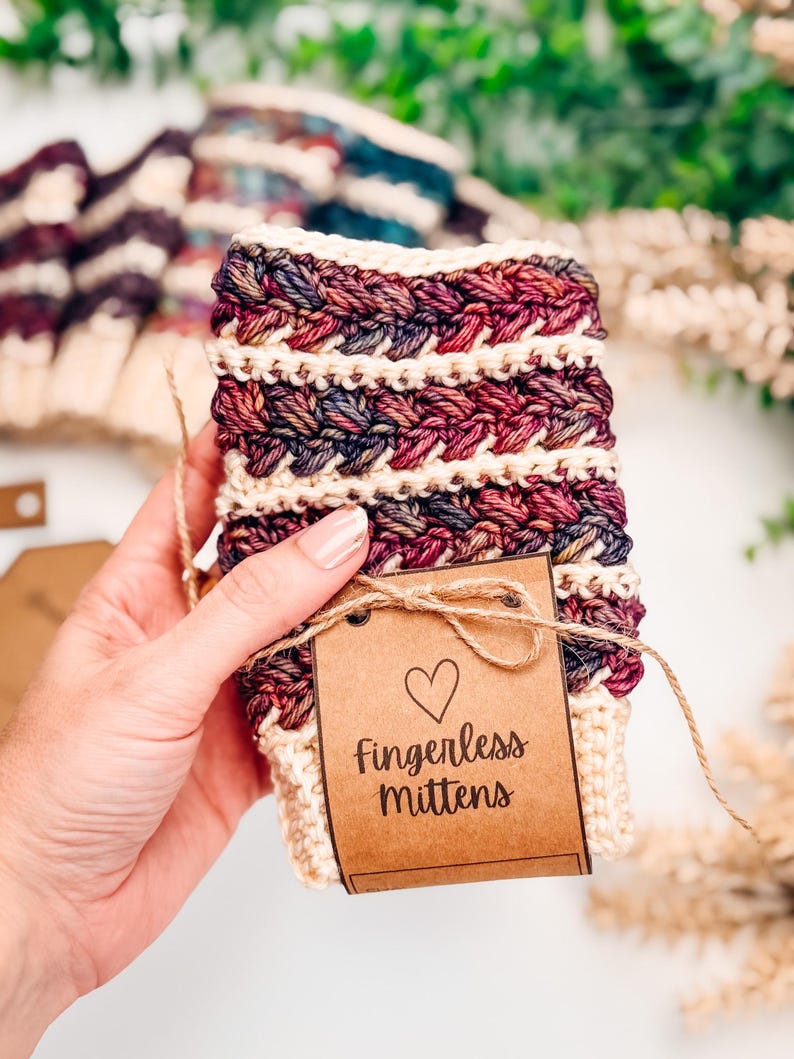 May include: Cream, burgundy, and purple fingerless mittens with a braided design. The mittens are handmade and have a brown tag that reads "Fingerless Mittens" with a heart symbol. The mittens are tied with twine.