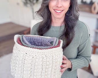 May include: Cream-colored, hand-crocheted basket with a textured cable-knit design. The basket has a round shape and contains three smaller, colorful crocheted baskets in shades of red, green, and purple. A cozy home decor item.